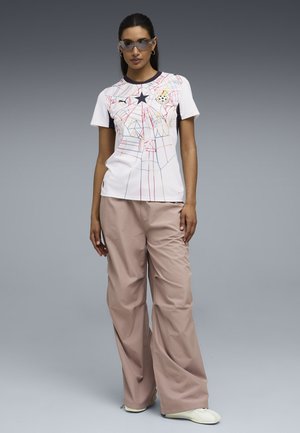 Kvinde står iført hvid T-shirt med grafisk mønster af geometriske linjer, beige bukser med vide ben, hvide sko og solbriller foran grå baggrund.