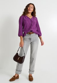 Blouse texturée violette avec un devant à boutons, jeans taille haute gris, mocassins en daim marron et un sac à main marron foncé.