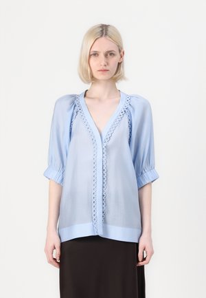 Blusa azzurra chiara con maniche a sbuffo e scollatura a V, con bordo ricamato lungo la parte anteriore e vestibilità ampia. Abbinata a una gonna scura.