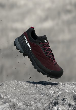 Μπορντό και μαύρο ορειβατικό παπούτσι Scarpa με ανθεκτική σόλα, αιωρούμενο πάνω από βραχώδη επιφάνεια με σωματίδια σκόνης.