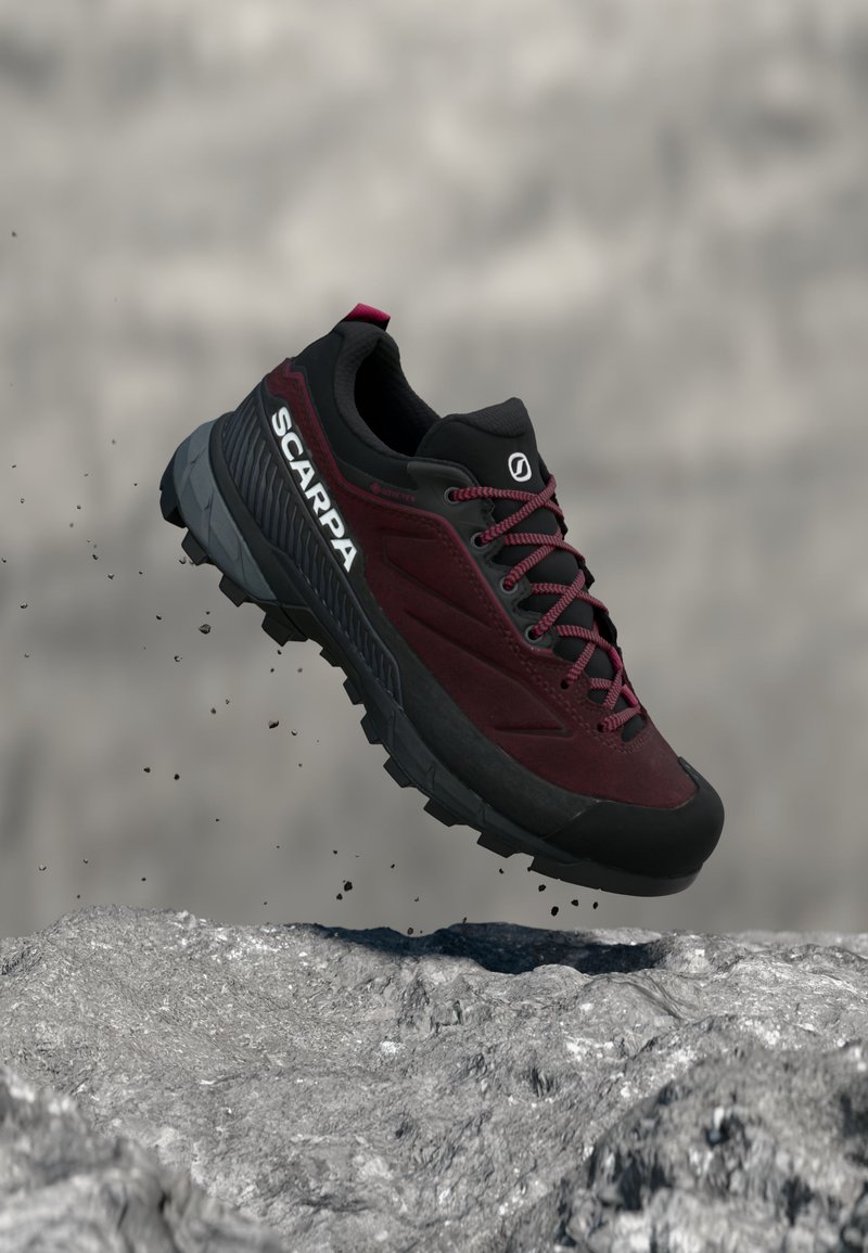 Chaussure de randonnée Scarpa bordeaux et noire avec semelle robuste, capturée en plein saut au-dessus d'une surface rocheuse avec des particules de poussière.