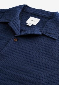 Marineblauwe polo met een textuur, voorzien van een knoopsluiting en een klassieke kraag. Etiket zichtbaar aan de binnenkant.