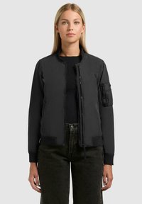 Donna che indossa una giacca bomber nera con zip e tasca sulla manica, abbinata a una maglietta nera e jeans scuri, in piedi davanti a uno sfondo semplice.