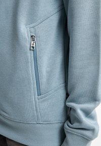 Ljusblå sweatshirt i mjukt material, med en ficka med dragkedja och ribbstickade muddar med synliga sömmar.