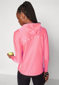 Felpa da performance rosa brillante con tessuto texture, maniche lunghe e grafica sul retro che recita "ENDLESS". Tiene una pallina da tennis in una mano.