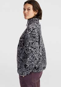 Pull-over en polaire noir et blanc à motifs, avec un col haut et une fermeture éclair, arborant des designs géométriques abstraits et une coupe décontractée.