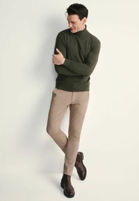 Ciemnozielony sweter z golfem,beżowe dopasowane spodnie, oraz brązowe botki za kostkę, na neutralnym tle. Gładki splot i dopasowany krój.