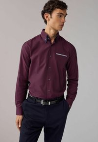 Camisa burdeos de botones con tejido texturizado, cuello oscuro en contraste y detalle del bolsillo con patrón, combinada con pantalones oscuros y cinturón.