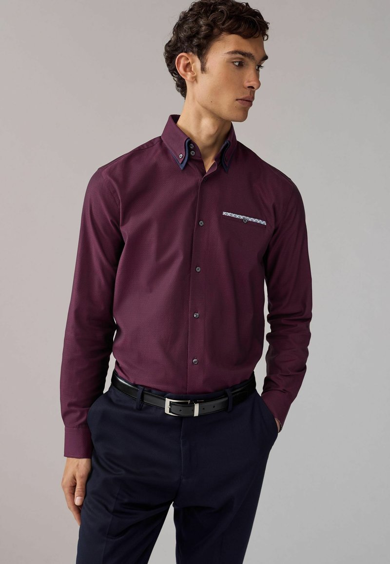 Camisa burdeos de botones con tejido texturizado, cuello oscuro en contraste y detalle del bolsillo con patrón, combinada con pantalones oscuros y cinturón.