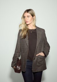 Blazer a quadri marrone in tessuto strutturato con grandi tasche, abbinato a una maglia scura, jeans scuri e una borsa marrone lucida.