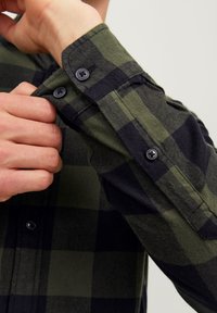 Grüner und schwarzer karierten Flanellhemdärmel mit zwei schwarzen Knöpfen und leicht strukturierter Stoff, wobei eine Hand die Manschette anpasst.