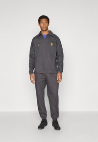 Pilkas megztinis su apykakle ir priekiu kišenėje, puoštas auksine siuvinėjimu, derinamas su atitinkančiomis siaurėjančiomis kelnėmis ir juodais sportbačiais.