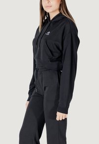 Felpa cropped nera con tasca frontale e coulisse. Abbinata a pantaloni jogging neri coordinati con vita e polsini elasticizzati.