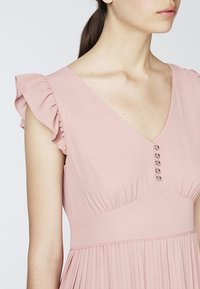 Frau in einem hellrosanen Kleid mit Rüschenshulterärmeln, einem V-Ausschnitt und fünf kleinen dekorativen Knöpfen auf der Brust.