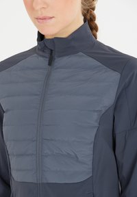 Kvinna som bär en tvåtonad mörkgrå jacka med en vadderad framdel och hög krage, flätat hår synligt bakifrån.