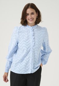 Blouse bleu clair à boutons avec col à volants et motif zigzag. Présente des manches bouffantes et des poignets élastiques. Portée avec un pantalon noir.