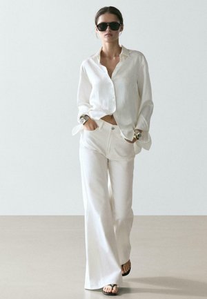 Donna che indossa una camicia bianca oversize con bottoni, pantaloni bianchi a gamba larga, occhiali da sole neri, infradito e accessori da polso, che cammina su uno sfondo semplice.