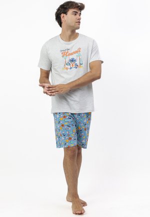 Joven descalzo que lleva una camiseta gris claro con el texto "Saludos desde Hawai" y un diseño de dibujos animados, combinado con pantalones cortos azules decorados con estampados coloridos.