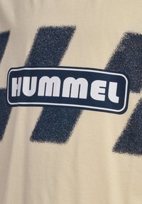 Beige stof med mørkeblå strukturerede diagonale striber og et afrundet rektangulært mærke med ordet "Hummel" i fede hvide bogstaver.
