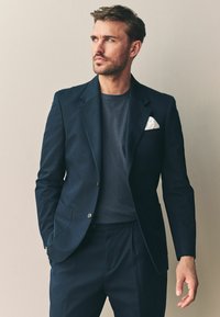 Marineblazer med en notch lapel, parret med en mørkegrå t-shirt og matchende bukser. Hvide lommetørklæde detaljer tilføjer kontrast.