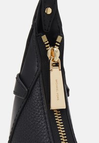Musta käsilaukku tekstuurilla varustetusta nahasta, jossa on kultainen vetoketju ja tunnus. Muotoilu sisältää jämäkän muodon ja sileät yksityiskohdat.
