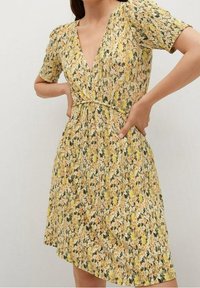 Vestido floral con un patrón amarillo y verde, mangas cortas, escote en V profundo, cintura ajustada y una tela que cae suavemente.