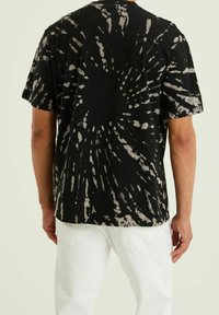 Muž v krátkém rukávu tričku s černo-bílým batikováním a bílých kalhotách, zobrazený zezadu na jednobarevném pozadí.