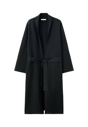 Žena s dlhými vlasmi, ktorá nosí dlhý čierny kabát, čierne nohavice a čierne topánky, stojí a pozerá sa cez rameno v minimalistickej miestnosti.