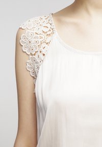 Haut sans manches en tissu crème clair avec bordure en dentelle florale sur l'épaule, présentant une texture lisse et une coupe ample.