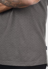 Chemise en tissu gris texturé avec un petit motif à pois, dotée d'une étiquette sur le côté. Le matériau paraît décontracté, avec une coupe confortable.