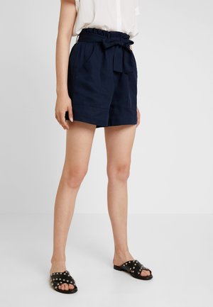 Shorts bleu marine avec une taille froncée, une ceinture à nouer et des poches latérales. Portés avec des sandales à lanières noires épaisses ornées de clous en argent.