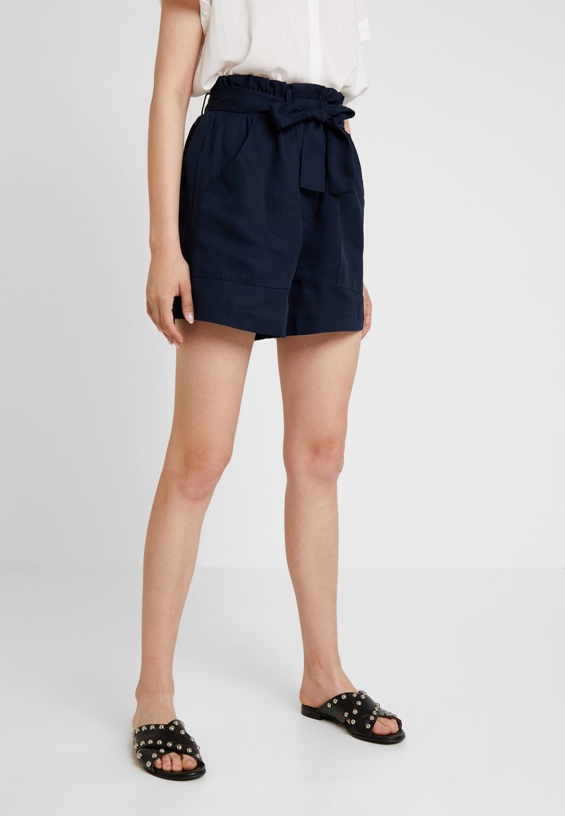 Námořnické modré šortky se staženým pasem, páskem na uvázání a bočními kapsami. Nosené s objemnými černými sandály na klínku s kovovými nýty.