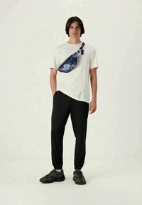 Hvite t-skjorte, svarte smale bukser og en blå mønstret crossover-veske. Modellen står mot en hvit bakgrunn, iført mørke joggesko.