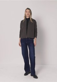 Veste grise zippée portée sur un col roulé noir, associée à un jean évasé bleu foncé. Le mannequin pose sur un fond blanc, portant des chaussures noires.