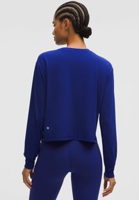 Bluză lungă cu mânecă, de culoare albastru royal, cu o croială lejeră, având un guler rotund și un detaliu mic cu logo la margine. Textură de țesătură netedă.