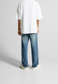 Vit oversized t-shirt med korta ärmar, kombinerad med lösa ljusblåa denimjeans och vita sneakers, sedd bakifrån.