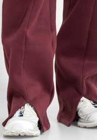 Bordeauxrote Sweatpants mit Seitenschlitzen am Saum, die weiße Sportschuhe zeigen. Weicher Stoff mit entspanntem Schnitt.