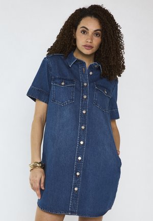 Femme aux cheveux bouclés portant une robe en jean à manches courtes avec des poches poitrine et des bracelets dorés au poignet gauche.