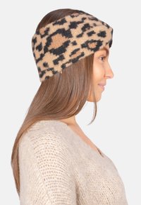 Bandeau en laine avec imprimé léopard en beige et noir. Design torsadé, largeur moyenne, texture douce. Porté sur de longs cheveux bruns.