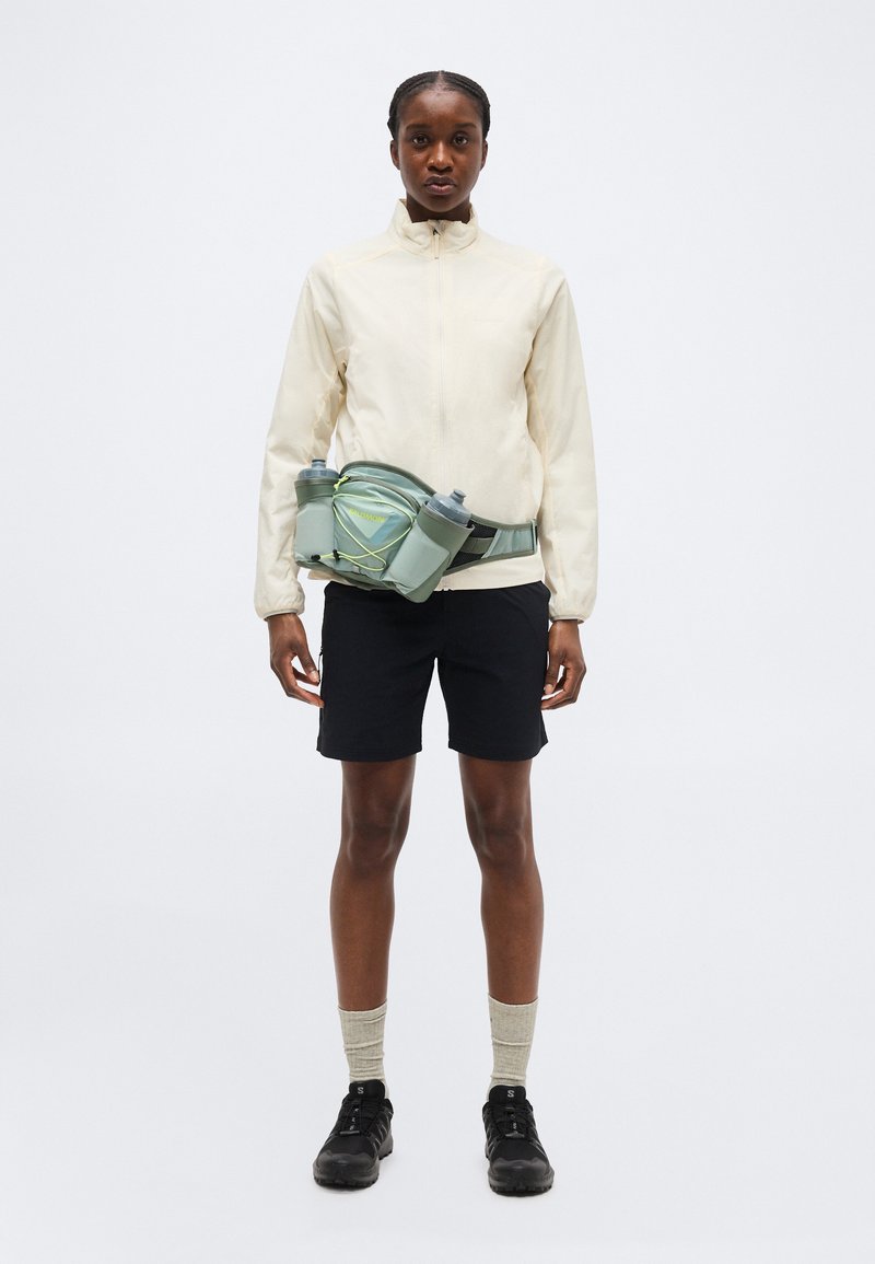 Personne portant une veste crème zippée, un short noir, des chaussettes beige, des chaussures noires et une poche ventrale verte claire avec deux bouteilles d'eau.