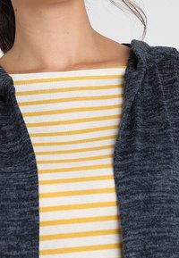 Mörkgrå huvtröja med mjuk textur, kombinerad med en krämfärgad och gul blus med horisontella ränder. Synlig halsringning och tygdetaljer.