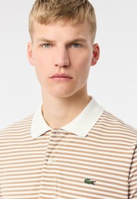 Beige och vit randig polo skjorta med en vit krage, som har en liten grön krokodillogo på vänster sida.