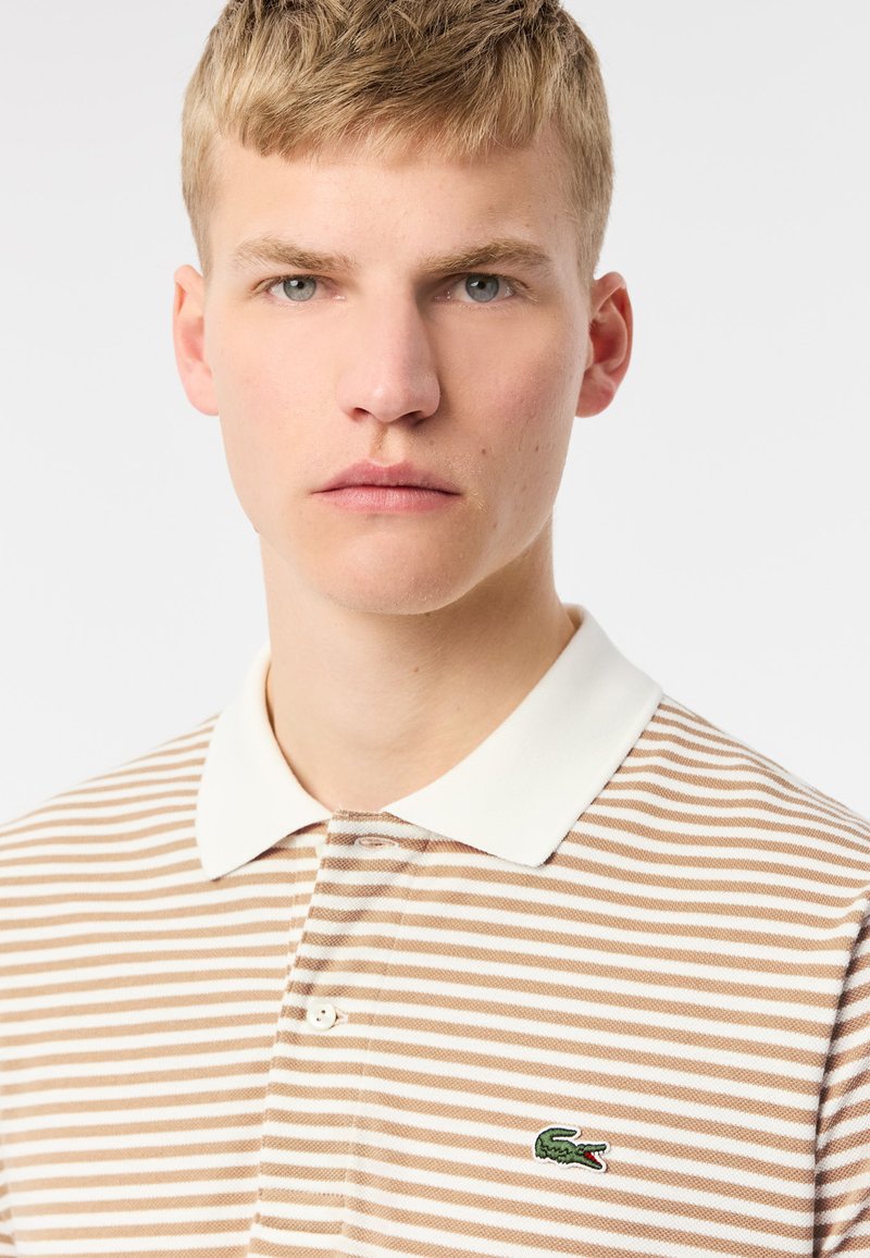 Polo shirt rayé beige et blanc avec un col blanc, portant un petit logo de crocodile vert sur le côté gauche.