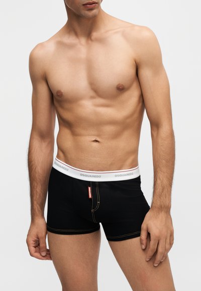 Torse masculin musclé portant un boxer noir avec une ceinture blanche portant l’étiquette DSQUARED2 sur un fond clair uni.