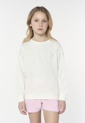 Ragazza bionda con occhi azzurri che indossa una felpa bianca a maniche lunghe con un piccolo logo rosa e pantaloncini rosa chiaro, in piedi su sfondo bianco.