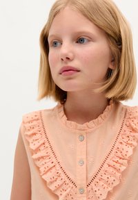 Pfirsichfarbener ärmelloser Bluse mit gerafftem Kragen, gesticktem Blumenmuster und Knopfverschluss. Leichter, atmungsaktiver Stoff.