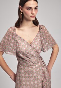 Wickelkleid aus leichtem Stoff mit einem pinken und grünen geometrischen Blumenmuster, tiefem V-Ausschnitt und flatternden Ärmeln. An der Taille gebunden.