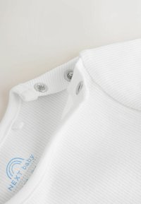 Bijela rebrasta dječja bodi majica s kopčama na ramenima. Oznaka je tiskana plavom bojom na unutrašnjosti. Mekani pamučni materijal, glatka tekstura.