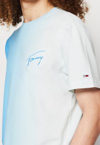 Svetle modré tričko s krátkými rukávy s gradientním přechodem, vybavené modře vyšitým logem "Tommy" na hrudi a malou značkovou nášivkou na rukávu.