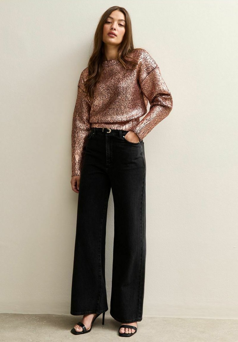 Suéter de manga corta en oro rosa con un acabado metálico texturizado, combinado con jeans de pierna ancha negros y sandalias de tacón alto negras con tiras.